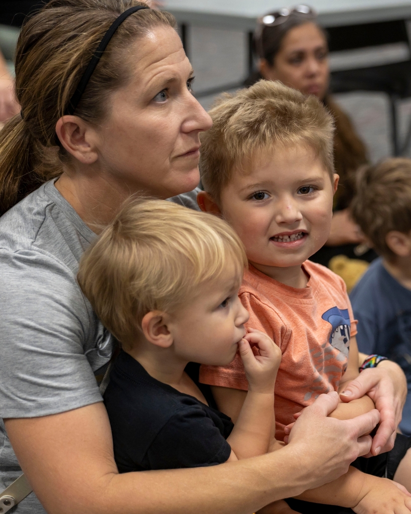 A woman sits on the floor holding two young children during a presentation.