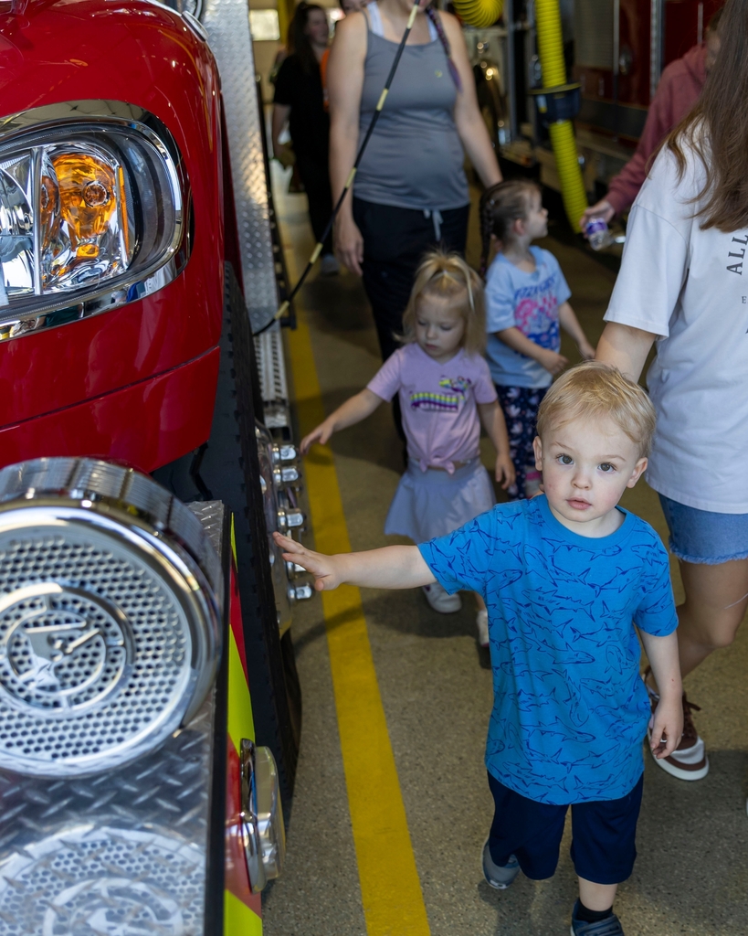 A toddler boy reaches out to touch the bumper of a red fire truck inside a fire station bay.