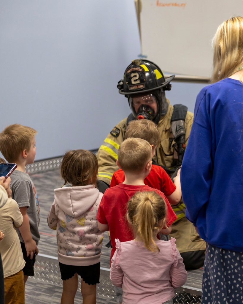 A group of young children gather around a firefighter kneeling in gear.