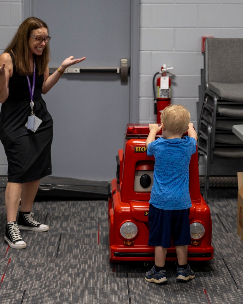 A young child stands next to a small red toy fire truck while an adult smiles in surprise.