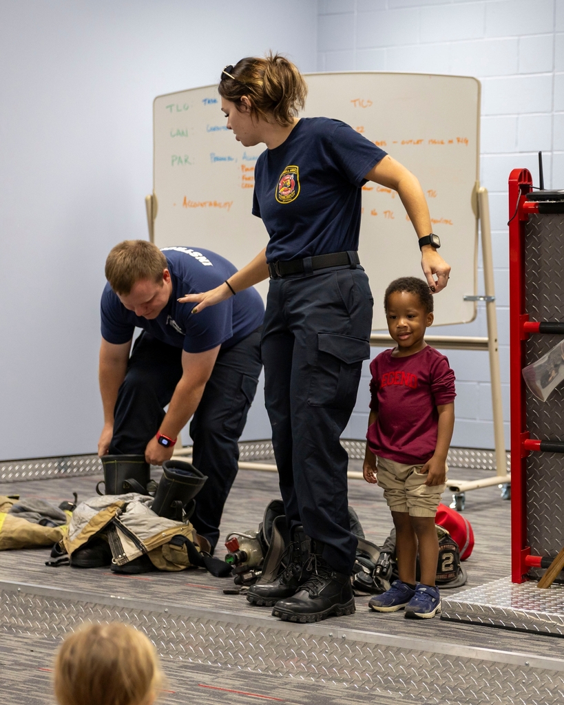 A female firefighter guides a young child on stage next to firefighter gear.