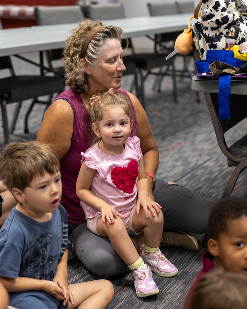 A toddler girl wearing a pink heart shirt sits on an adult's lap on the floor listening attentively.