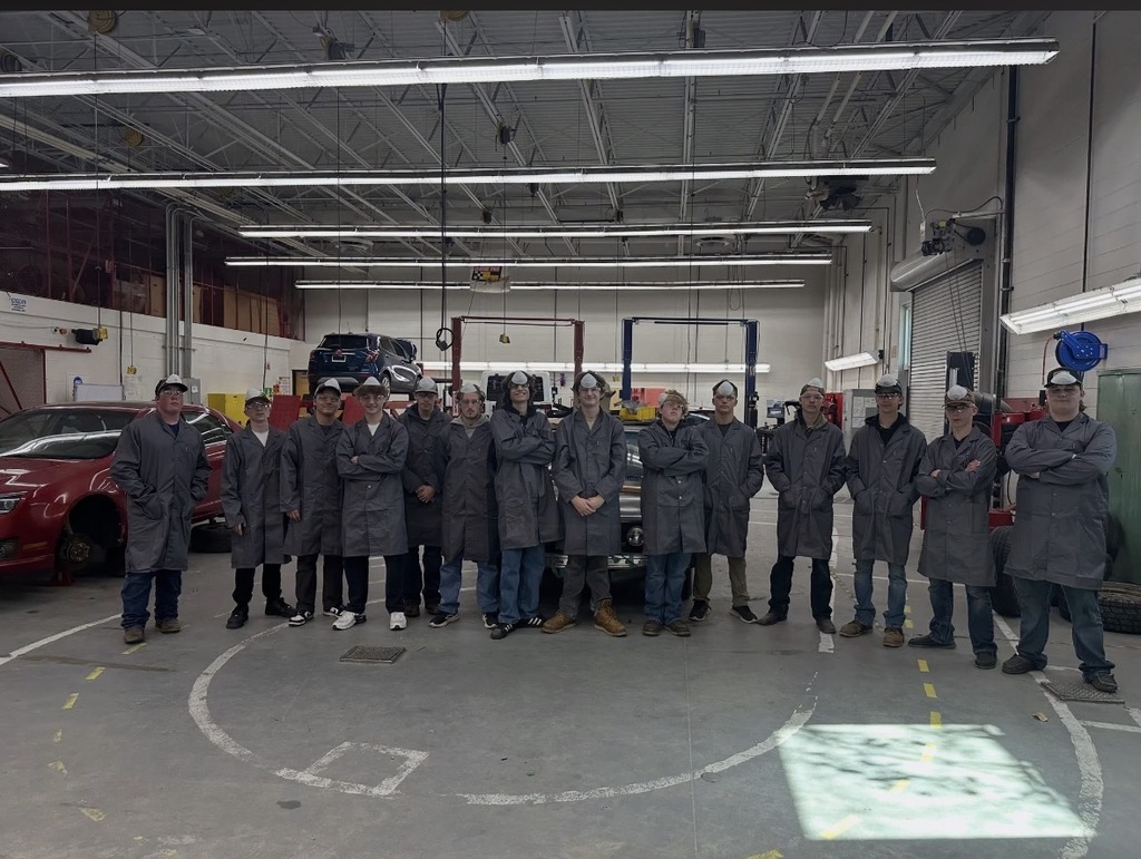 A group of students wearing gray automotive shop coats and safety glasses pose for a group photo inside an automotive technology training shop, with vehicles and car lifts in the background.