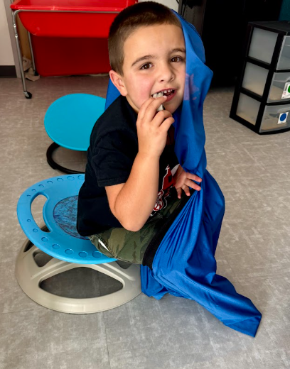 A young boy smiles while sitting on a blue balance stool with a stretchy blue sensory sock draped over his head and shoulder.