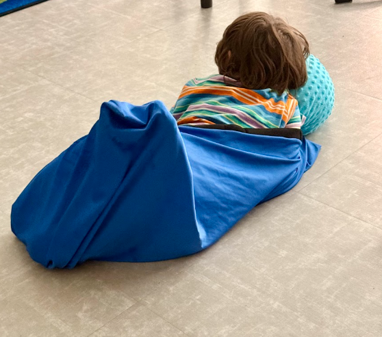 A young child wearing a colorful striped shirt lies on the floor inside a stretchy blue sensory body sock, resting their head on a textured light blue pillow.