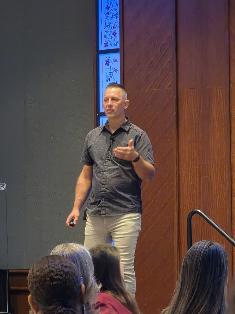 Speaker Ted Kroll presents on stage, gesturing with his left hand while holding a slide clicker in his right. He is wearing a dark short-sleeve button-up shirt and khaki pants, viewed from behind the audience.
