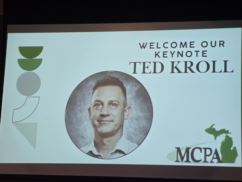A presentation title slide displaying "WELCOME OUR KEYNOTE TED KROLL" above a circular photo portrait of Ted Kroll. Abstract geometric shapes sit on the left, and an MCPA logo with a green Michigan state map outline is in the lower right corner.
