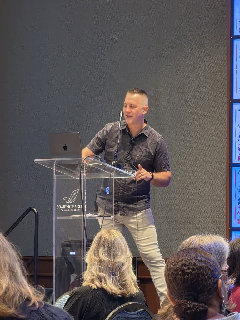 Keynote speaker Ted Kroll stands behind a clear podium labeled "Soaring Eagle Casino & Resort," gesturing while speaking with an open laptop on the stand in front of him. He wears a dark short-sleeve button-down shirt and light pants, pictured from behind audience members