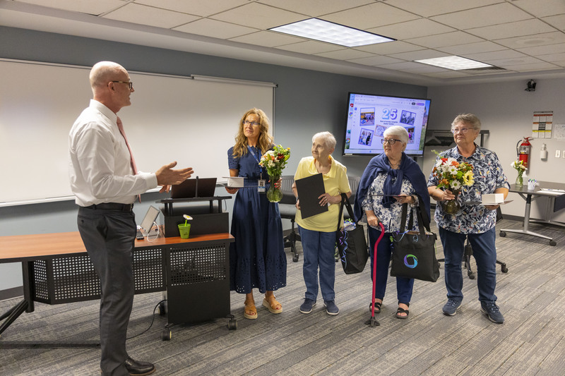 A speaker in a dress shirt addresses four women holding flower bouquets and certificates in a conference room.
