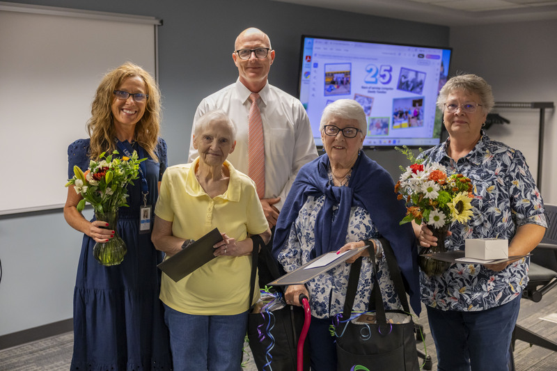 Five people stand together in a meeting room holding flowers and awards for a group photo.
