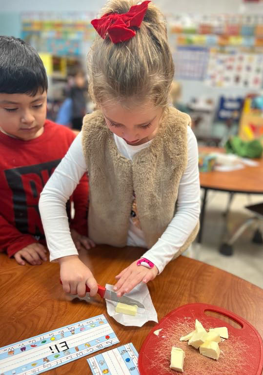 A student uses a knife to cut butter as another student watches