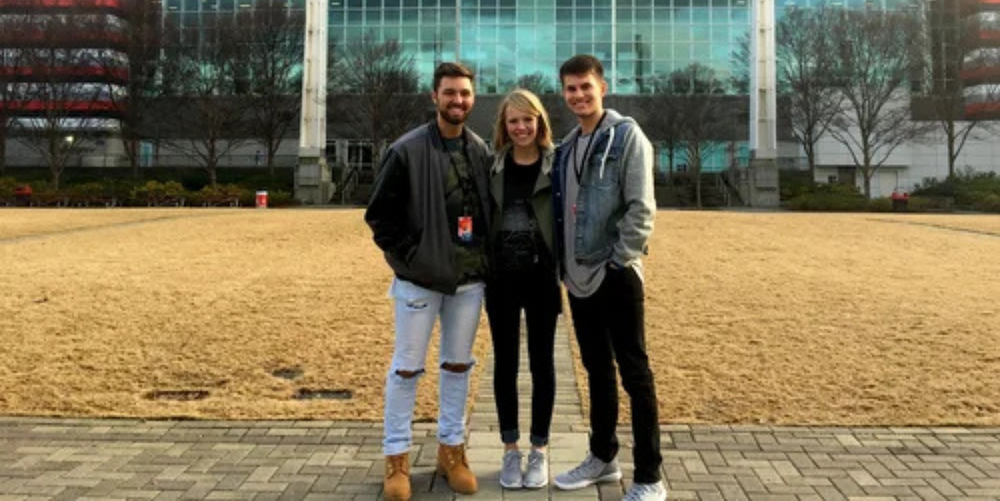 Two black haired high school aged boys stand on both sides of a blonde high school aged girl while posing for a photo in front of a grass field and a glass building behind them.