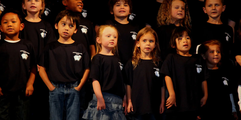 A group of young children stand on a stage wearing black T-shirts while singing during a school program.