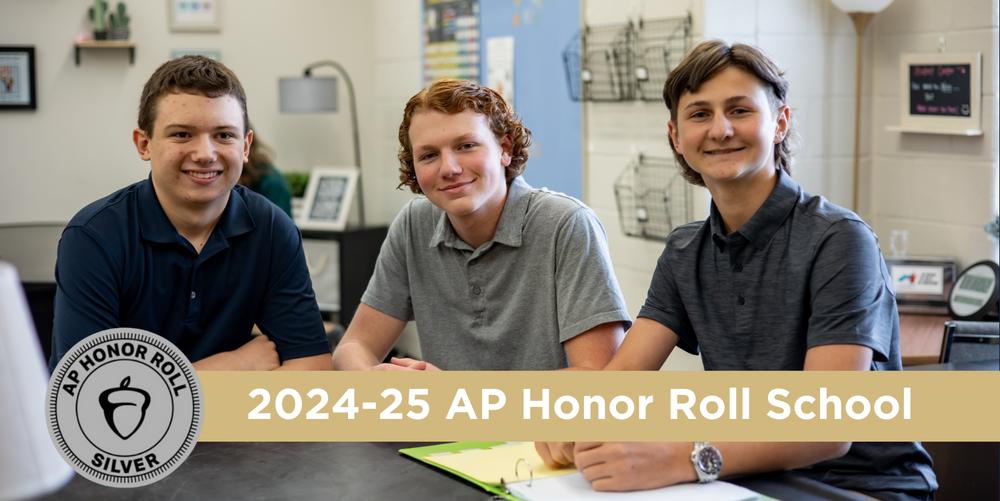 Three dark haired boys sit at a desk wearing dark colored shirt. They're smiling, posing for a photo with their hands folded in front of them.