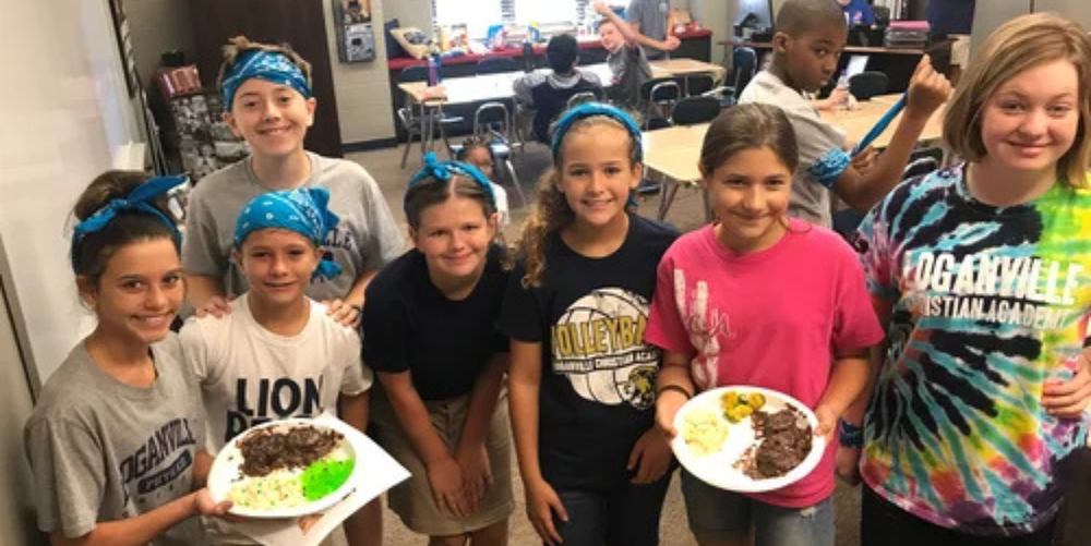 Seven middle school aged students pose smiling for a photo inside a classroom showing off their school project.