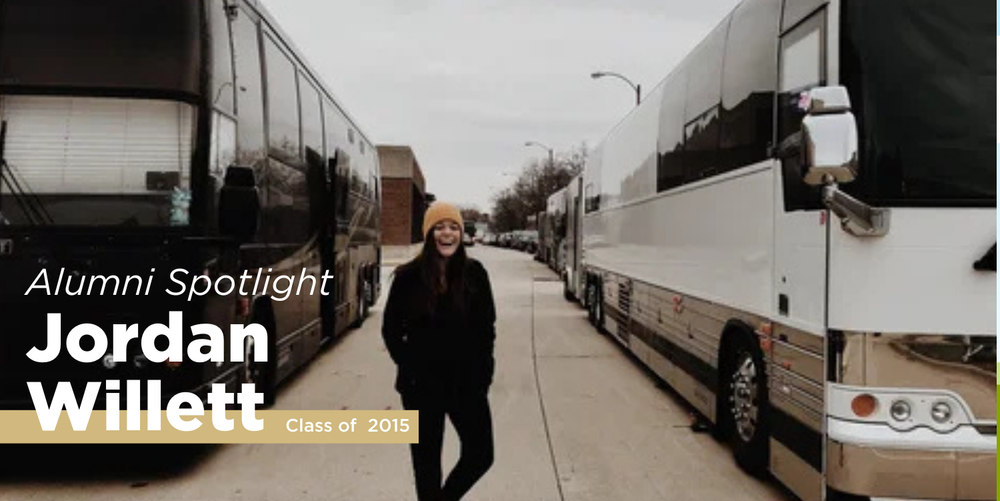 A black haired woman wearing a yellow winter hat standing in between a black bus and a white bus poses for a photo smiling.