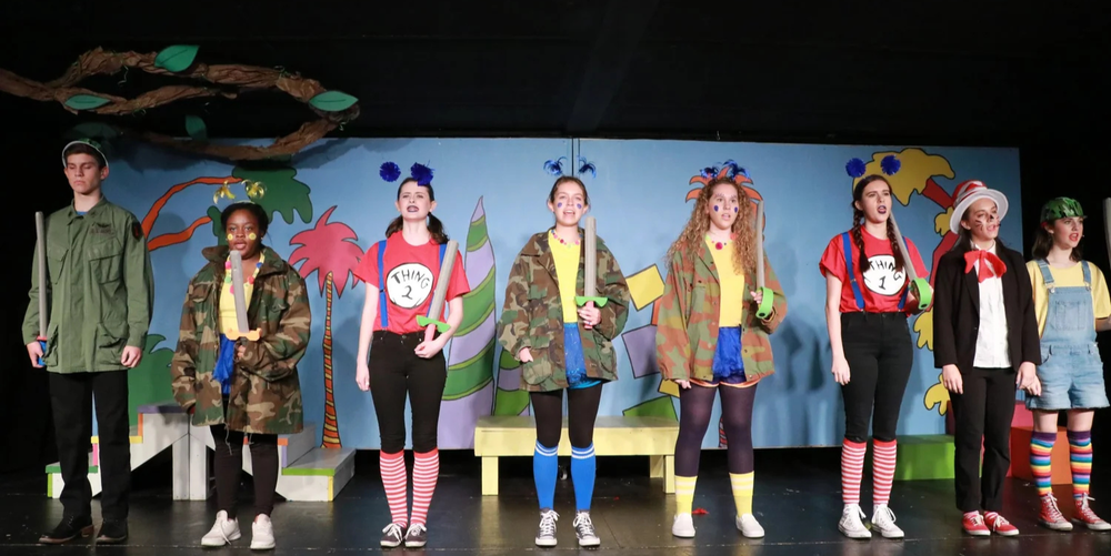 A group of students stand in one line while performing  a play in a black box theater.