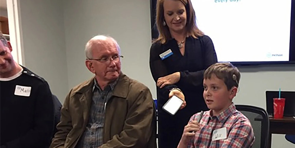 A older man sits to the left of a young boy while they speak to a crowd of people. 