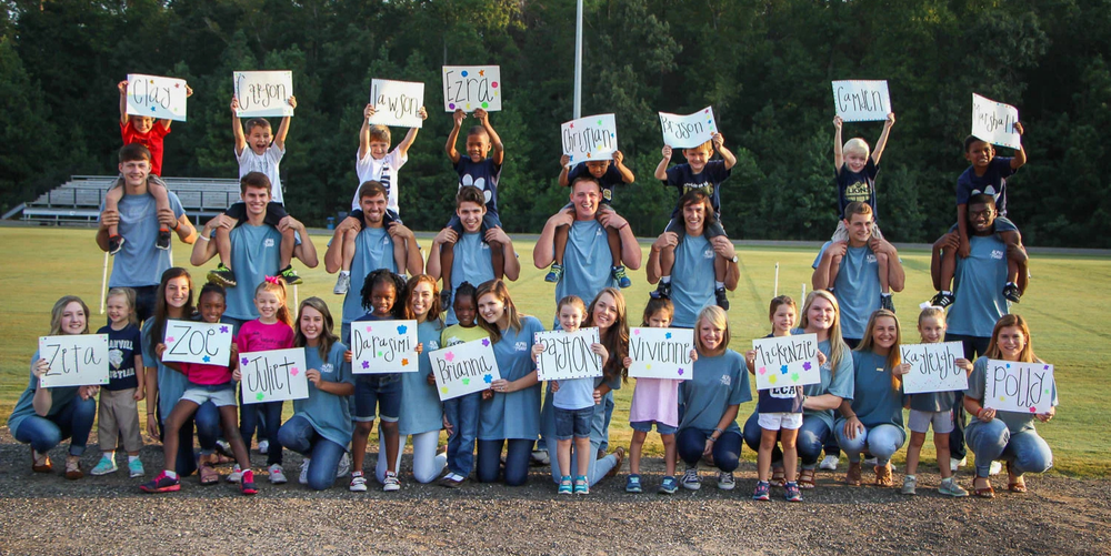 A group of high school aged students pose for a group photo with younger children while holding white decorative signs and smiling. 