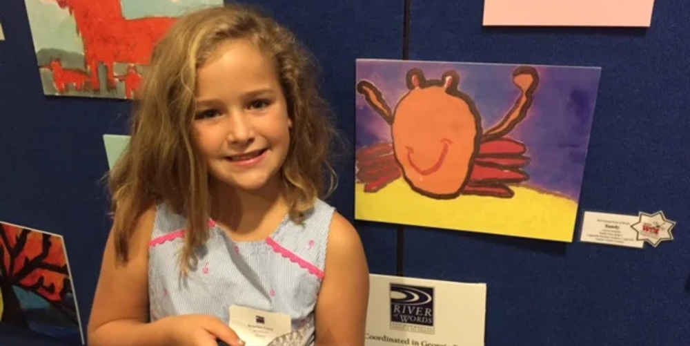 A young lower school aged blonde haired girl smiles and poses for a photo next to an art design hanging on the wall next to her.