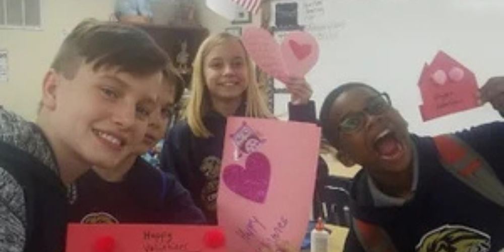 Four students pose smiling for a photo while holding up bright pink paper designed for Valentines Day. 