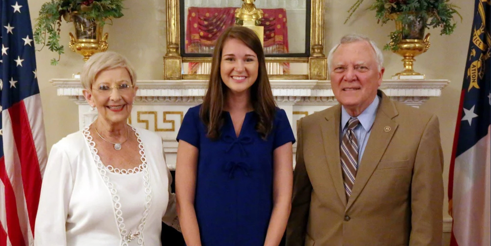 A young brown haired girl wearing a blue dress while smiling and posing for a photo with one older woman and one older man dressed nicely..