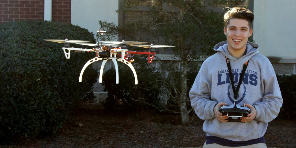 A brown haired high school aged boy wearing a light grey and dark blue hoodie holds a remote control while his drone hovers next to him.