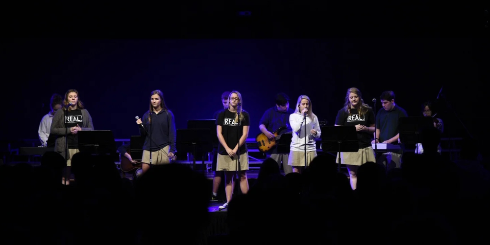 A group of five students stand at the front of a dark stage in a dark room preparing to lead worship at a Chapel service.