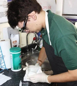 A black haired teenage boy wearing a green shirt and clear goggles, looks over a clear contained in a science lab. 