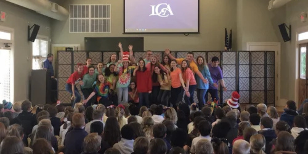 A large group of students wearing bright colored shirts make a presentation in front of an even larger group of people. 