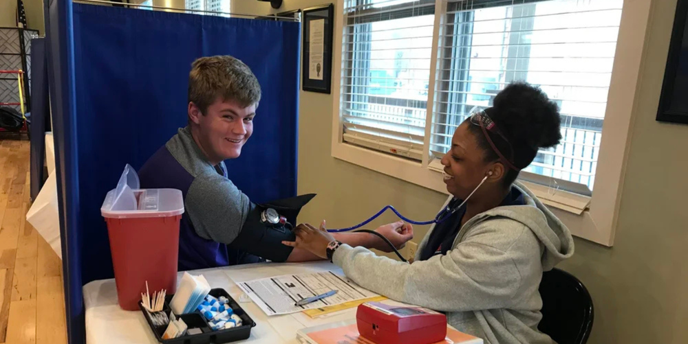 A brown haired boy smiles while a nurse takes his blood pressure ahead of donating blood. Both are laughing in the photo.