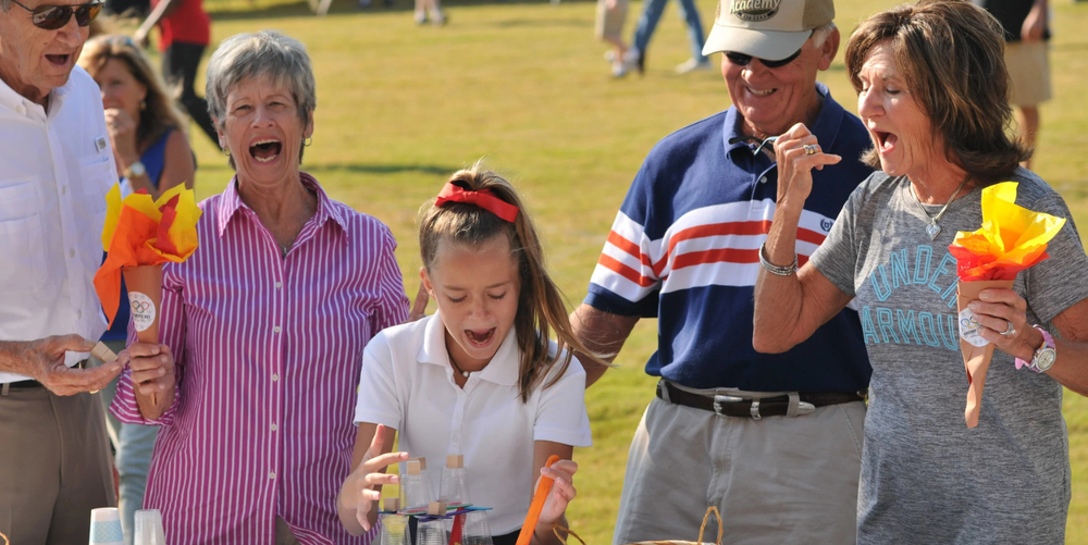 Four older people stand around a young girl smiling and laughing as their grandchild plays a game outside in a grass field. 