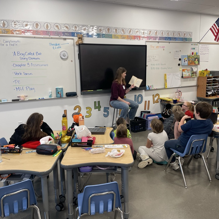 A woman reads aloud to a class, who is sitting in desks and on the floor 