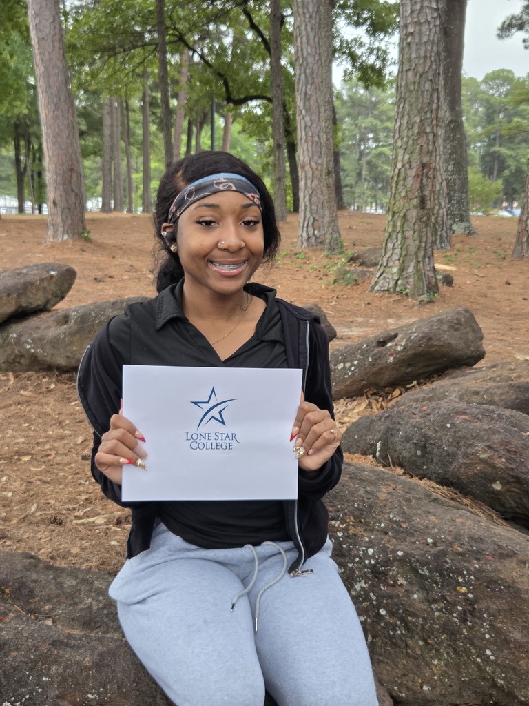 Aarianna holding a Lonestar College sign and smiling while sitting on a large rock surrounded by trees.