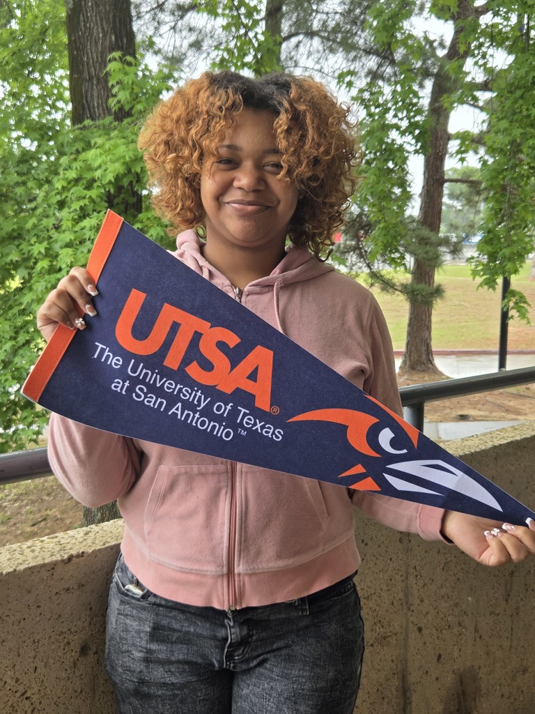 Kyla is standing outside holding  a UTSA pennant with a roadrunner head and trees in the background.