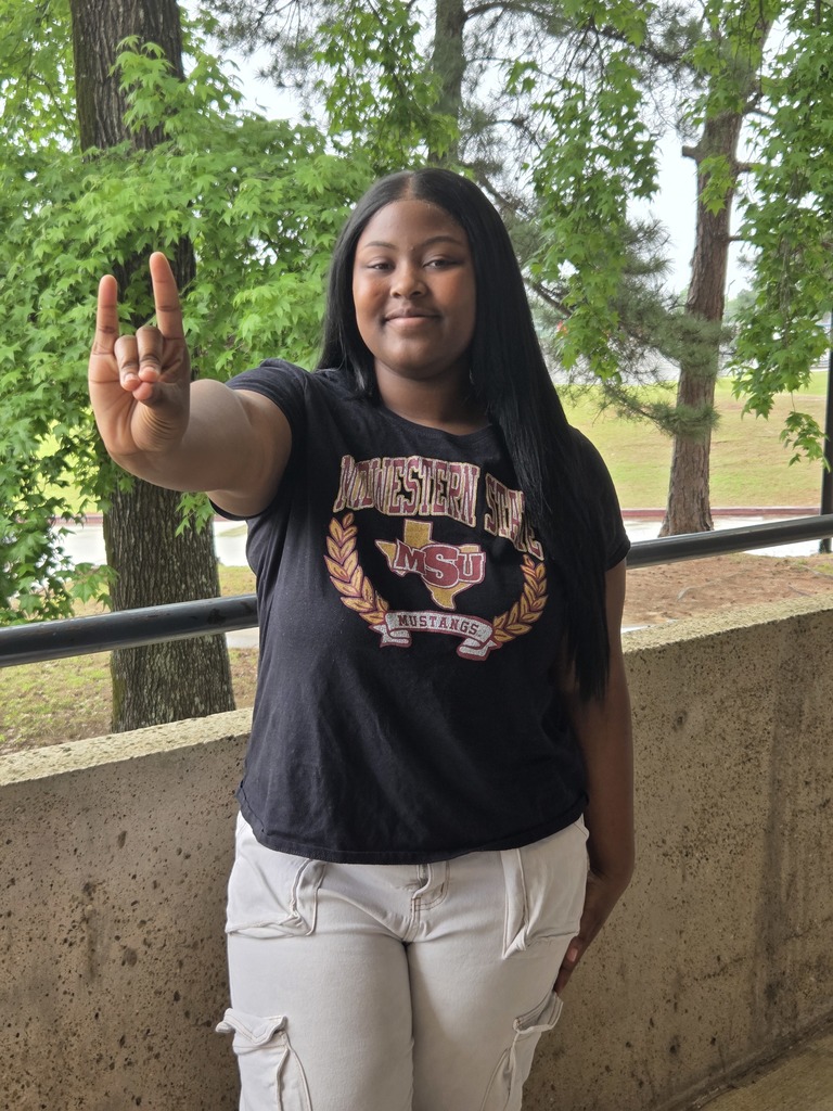 Alycea standing outside with trees in the background wearing her Midwestern State University shirt and holding her hand out presenting the mustang hand sign.