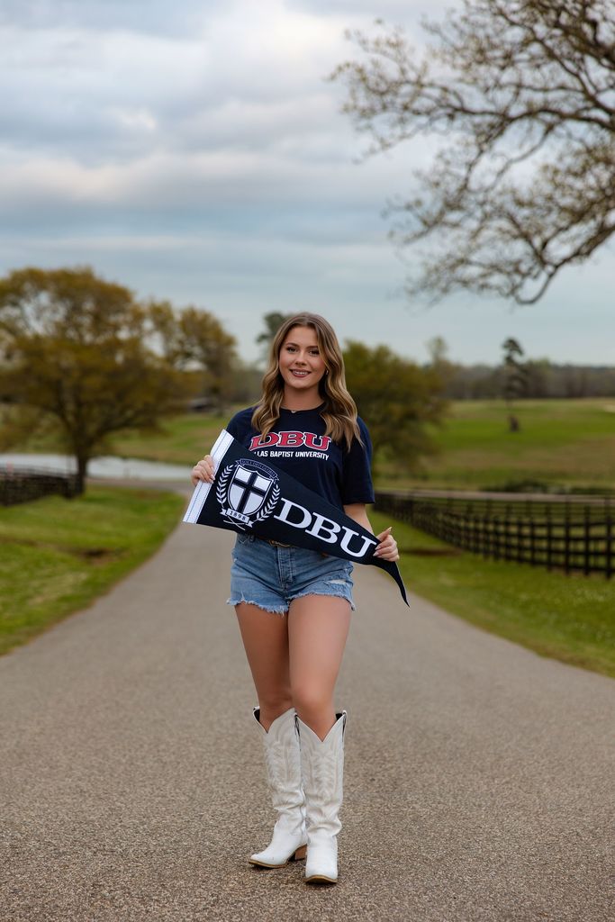 Bella standing on a road with farm land around her holding a DBU pennant and wearing a DBU shirt