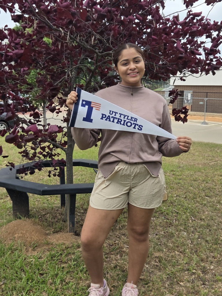 Zoraya is standing in front of a tree with purple leaves holding a UT Tyler Patriots pennant.