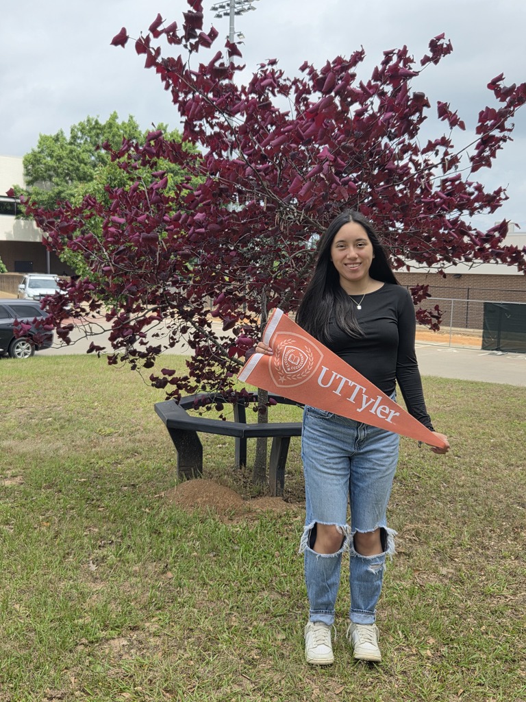 Itzel is holding a UT Tyler pennant standing in front of a tree with purple leaves.