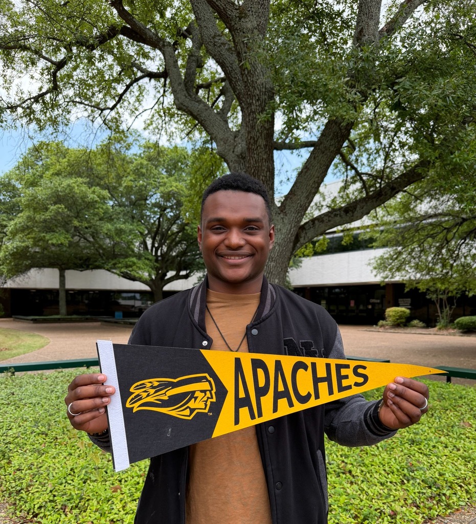 Chris is outside with a tree in the background holing a Tyler Junior College pennant in the background.