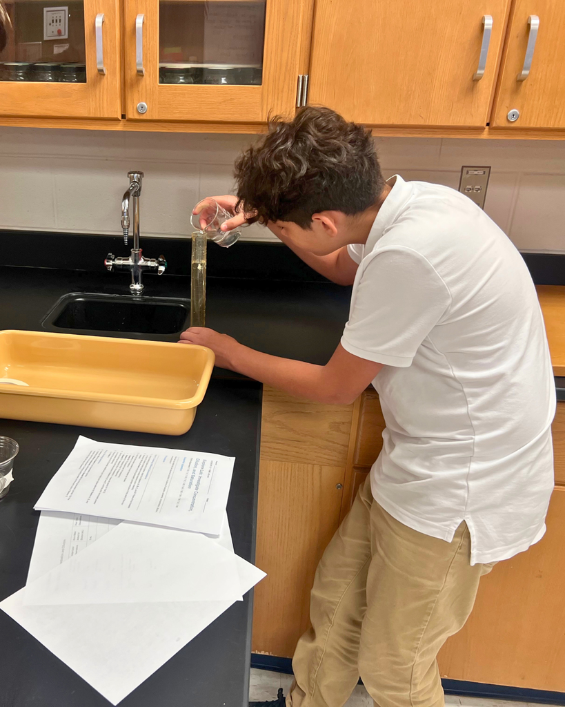 A student leaning over a lab table to carefully pour liquid into a graduated cylinder during a science experiment.