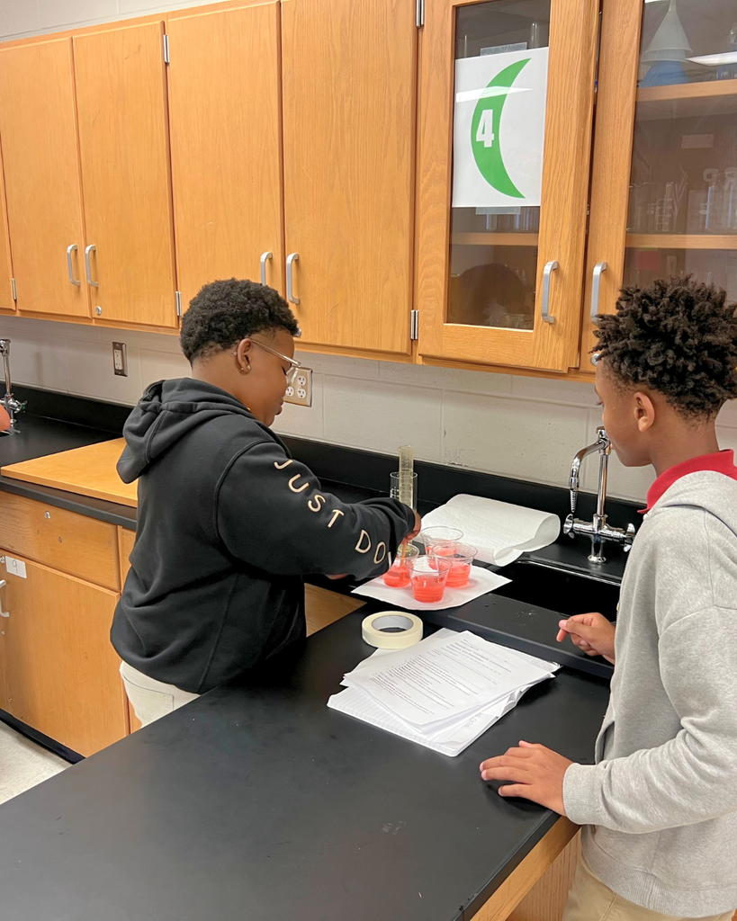 A student leaning over a lab table to carefully pour liquid into a graduated cylinder during a science experiment.