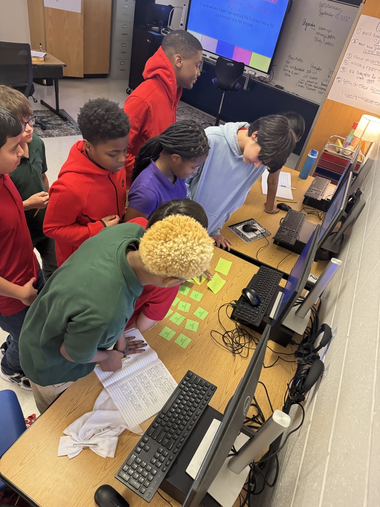 Middle school students standing around a classroom computer desk, examining bright green sticky notes and papers placed near the keyboard and monitor.