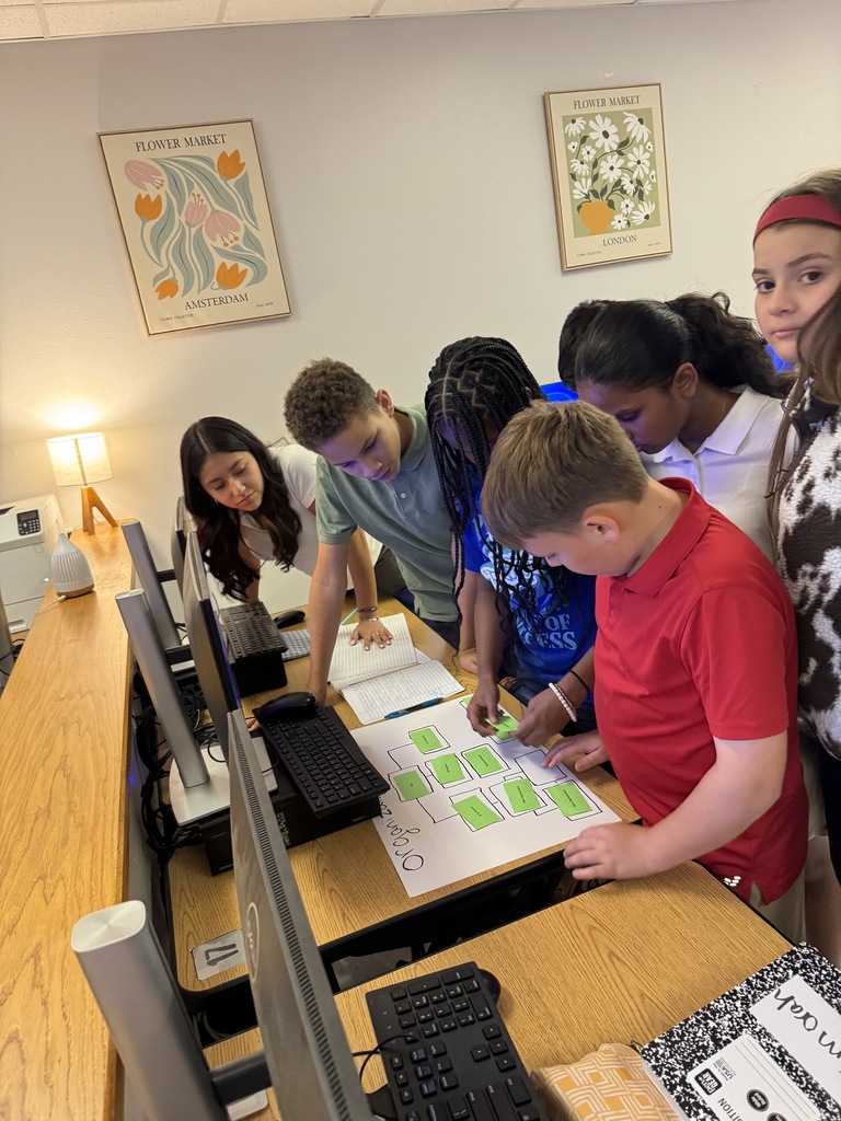 Middle school students leaning over a classroom computer desk to work together on an activity with green sticky notes and paper worksheets.
