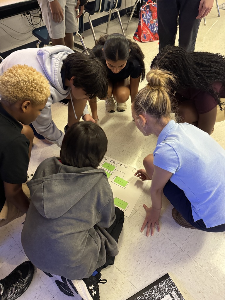 Middle school students gathered closely on the classroom floor around green sticky notes and papers for a collaborative activity.
