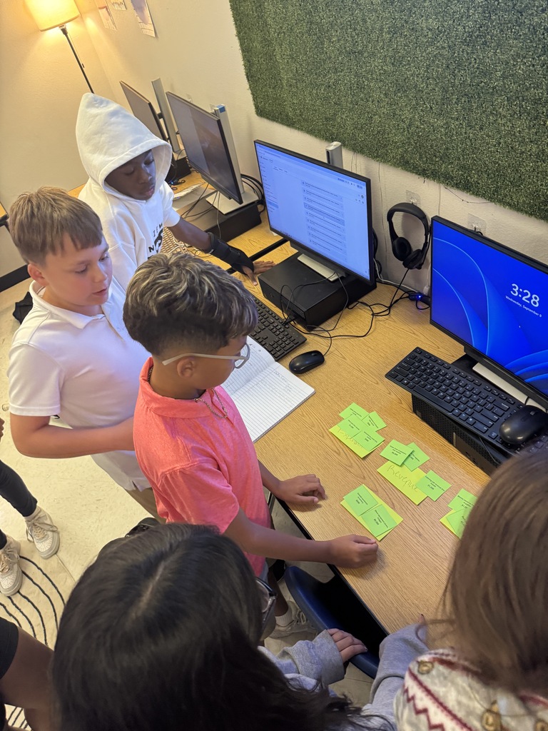 iddle school students gathering around a classroom desk with computers and green sticky notes to collaborate on a hands-on activity.