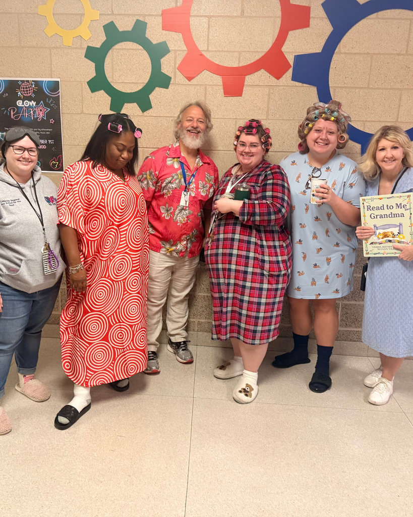 A group of six school staff members dressed in fun costumes posing together in a hallway in front of wall decorations.