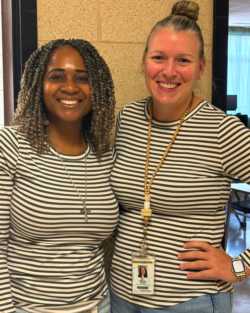 Two smiling women wearing matching black-and-white striped shirts standing side-by-side indoors.