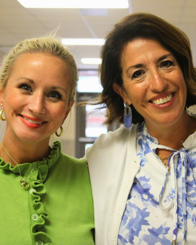 Two smiling women standing side-by-side indoors, one wearing a green high-neck top and the other wearing a blue-and-white patterned top with a light cardigan.