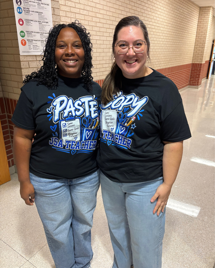 Two smiling women standing together in a school hallway wearing matching black "PASTE / COPY" teacher t-shirts and jeans.